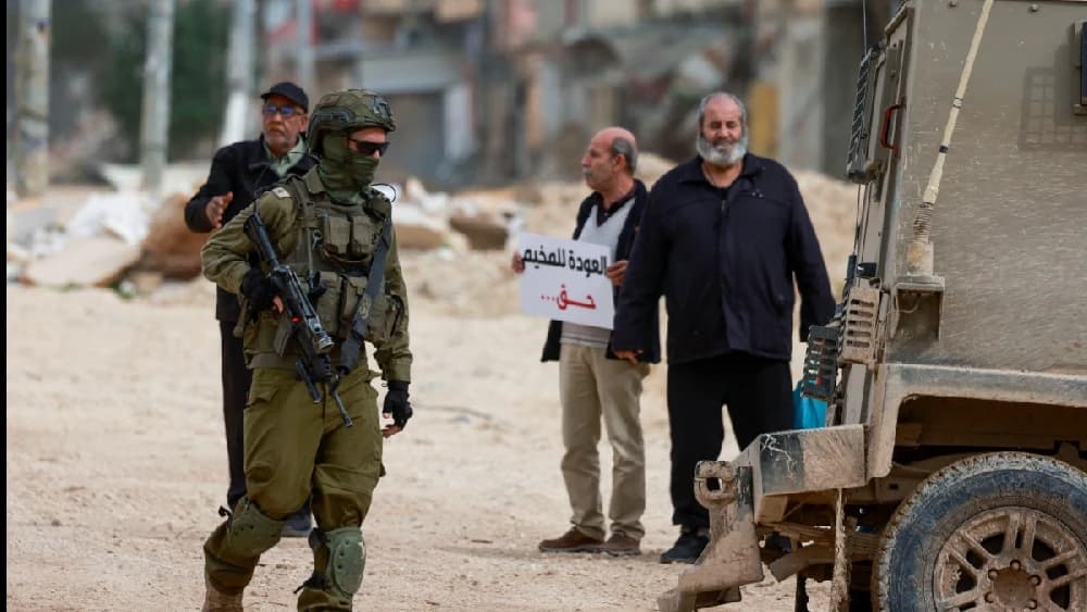 An Israeli soldier walks near Palestinians protesting after Israeli security forces blocked the entrance of the Nur Shams Palestinian refugee camp in the occupied West Bank on February 9, 2026, demanding to be allowed to return to their homes from where t